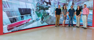five people standing in front of a large screen showing a pellet mill