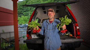 A grower holding fresh radishes in each hand while standing in front of his open pickup truck bed at a farmers market